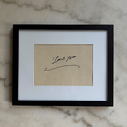 A framed art print featuring a beige textured card with a black cursive signature that reads 'I Love You', set in a white mat and a black frame, resting on a light marble surface.