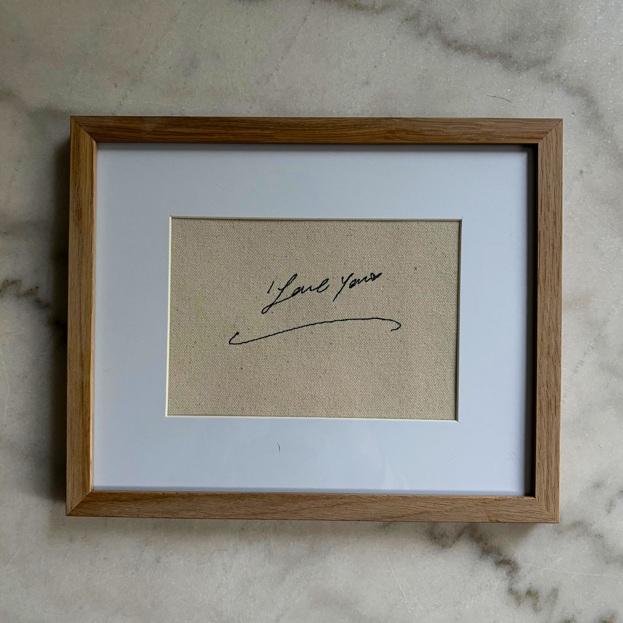 A square framed art print featuring a beige textured background with a black cursive signature that reads 'I love you', set within a white mat and a light brown wooden frame, resting on a light gray marble surface.
