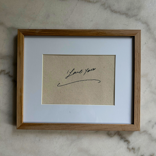 A square framed art print featuring a beige textured background with a black cursive signature that reads 'I love you', set within a white mat and a light brown wooden frame, resting on a light gray marble surface.