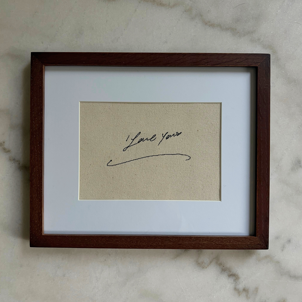 A square framed art print featuring a beige textured background with a black cursive signature that reads 'I love you', set within a white mat and a dark brown wooden frame, resting on a light gray marble surface.
