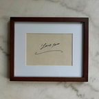 A square framed art print featuring a beige textured background with a black cursive signature that reads 'I love you', set within a white mat and a dark brown wooden frame, resting on a light gray marble surface.