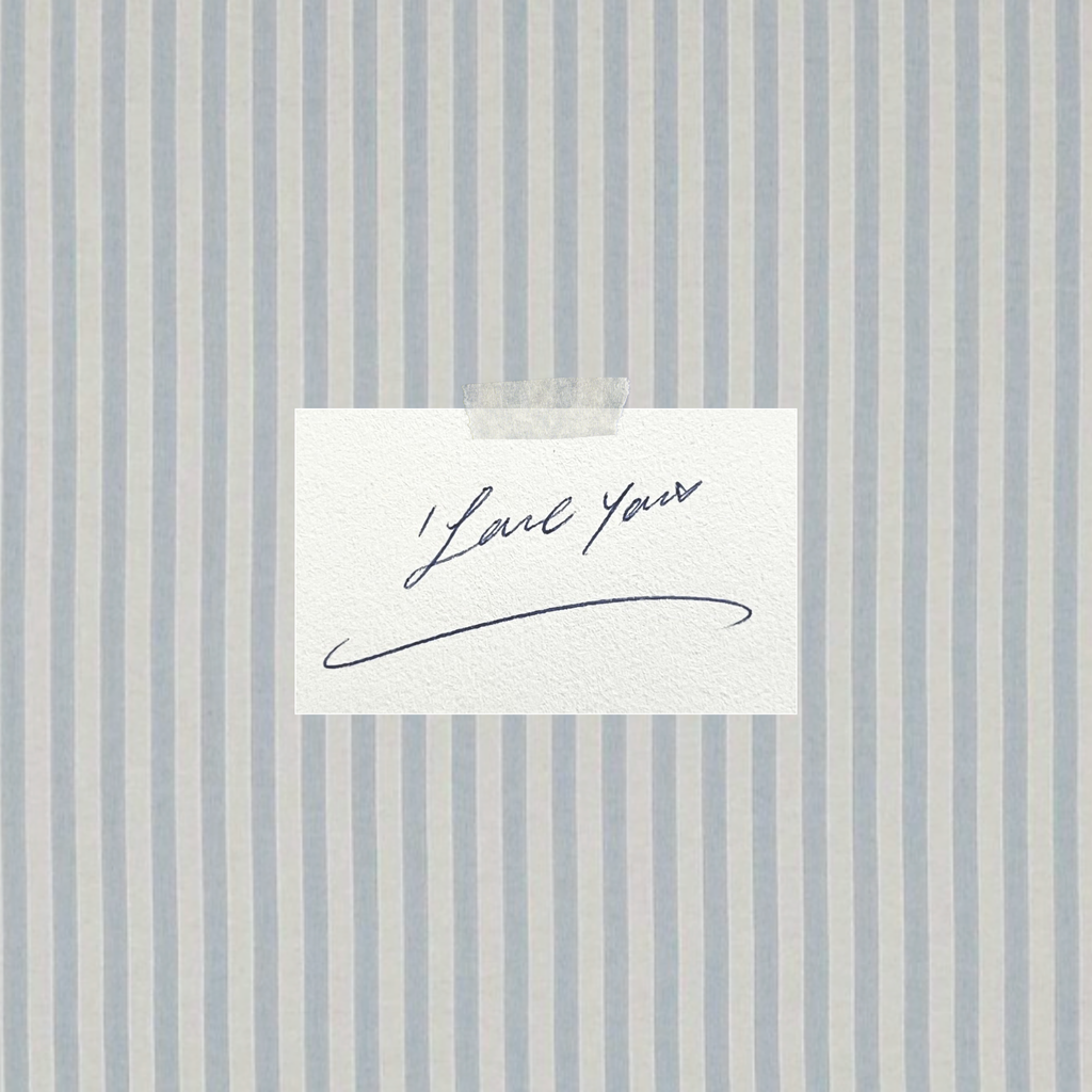 A small white card with a handwritten script reading 'Love You' taped against a light blue and white vertically striped background.